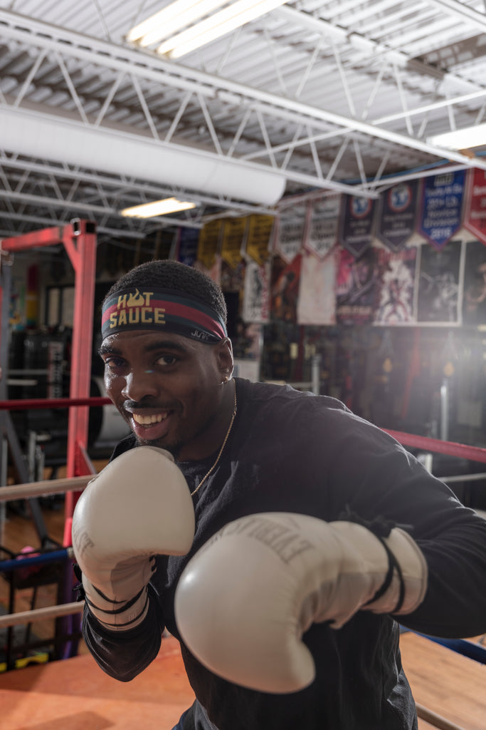 This image shows Kalvin, a black man, in his white boxing gloves, black long sleeved t shirt, and hot sauce junk headband. He is shadowboxing the camera. He is in the fighting ring and there are posters and banners behind him on the walls. 