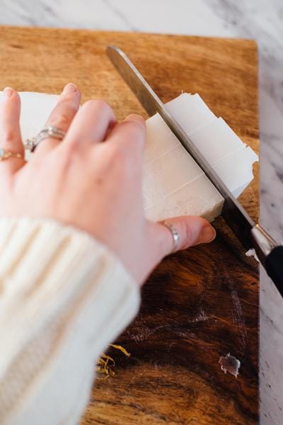 Hand and knife cutting soap base bar into cubes on wooden cutting board.