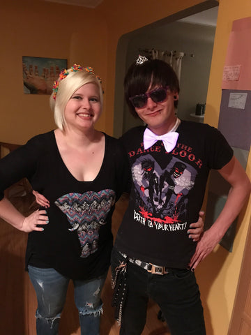 Couple wearing emo-goth outfits with the blond woman wearing a rainbow flower crown and the dark haired man wearing a tiny tiara and light up fiber optic bow tie