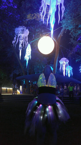 woman with her head thrown back and arms out wearing a light up tulle skirt and fiber optic cropped halter top under lit up sculptures hanging from trees in a dark forest with other festival goers in the background