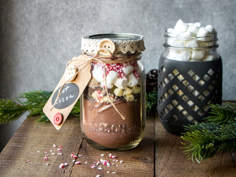 Two Mason jars are filled with the ingredients to make hot cocoa, including marshmallows and peppermint.