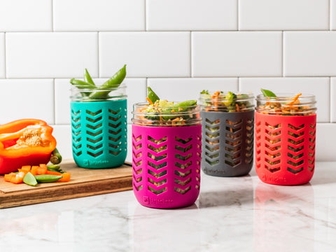 4 16oz wide mouth mason jars fitted with JarJackets silicone sleeves sit on a kitchen countertop. A wooden cutting board sits beneath one jar. Chopped red pepper and snap peas also sits on the cutting board. All 4 jars are filled with a farro salad and vegetables.