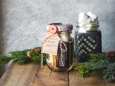 A mason jar filled with Hershey’s bars, marshmallows and graham crackers and decorated with a gift tag and festive twine.