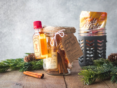 Two Mason jars are filled with all of the ingredients needed to make spiked apple cider, including a travel-sized bottle of Fireball Cinnamon Whiskey.