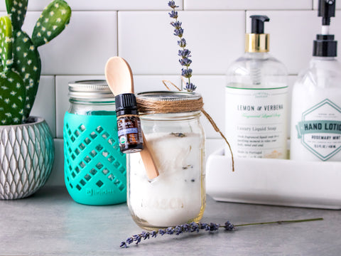 Two Mason jars are full of a body scrub made with lavender buds. One has a small wooden spoon and bottle of lavender essential oil attached to it with twine. 