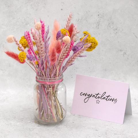Pastel pink dried flowers in a glass vase next to a pink congratulations greeting card