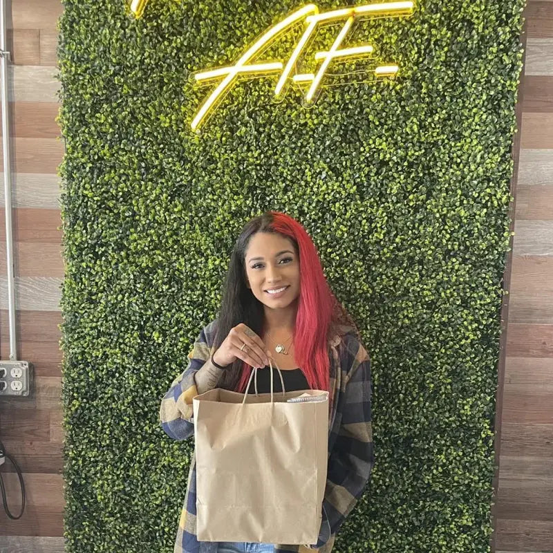 Smiling woman holding a brown paper bag in front of a green leafy wall with neon writing.