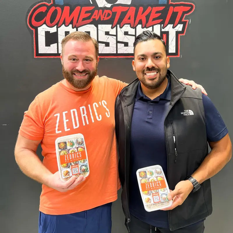 Two men holding Zedric's meal packages, standing in front of a 'COME AND TAKE IT CROSSFIT' sign.