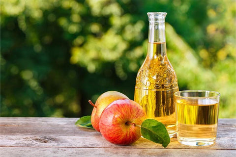 A glass bottle and a glass of apple cider vinegar alongside 2 apples on a table with nature in the background