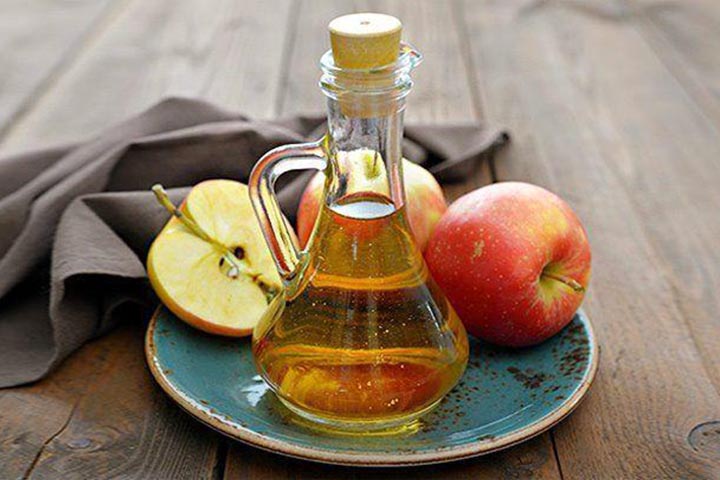 A glass bottle of apple cider vinegar next to 3 apples on a plate next to a cloth