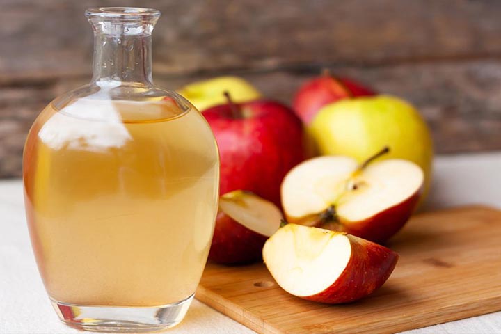 A full glass jar of apple cider vinegar next to red and yellow apples on a wooden cutting board