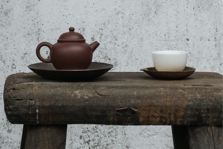 A tea kettle and a white coloured cup of tea on a wooden stool