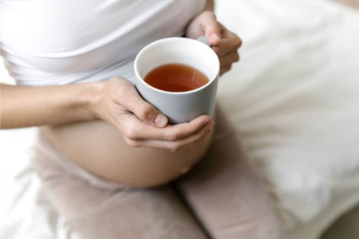 A pregnant woman in white top holding a cup of raspberry leaf tea