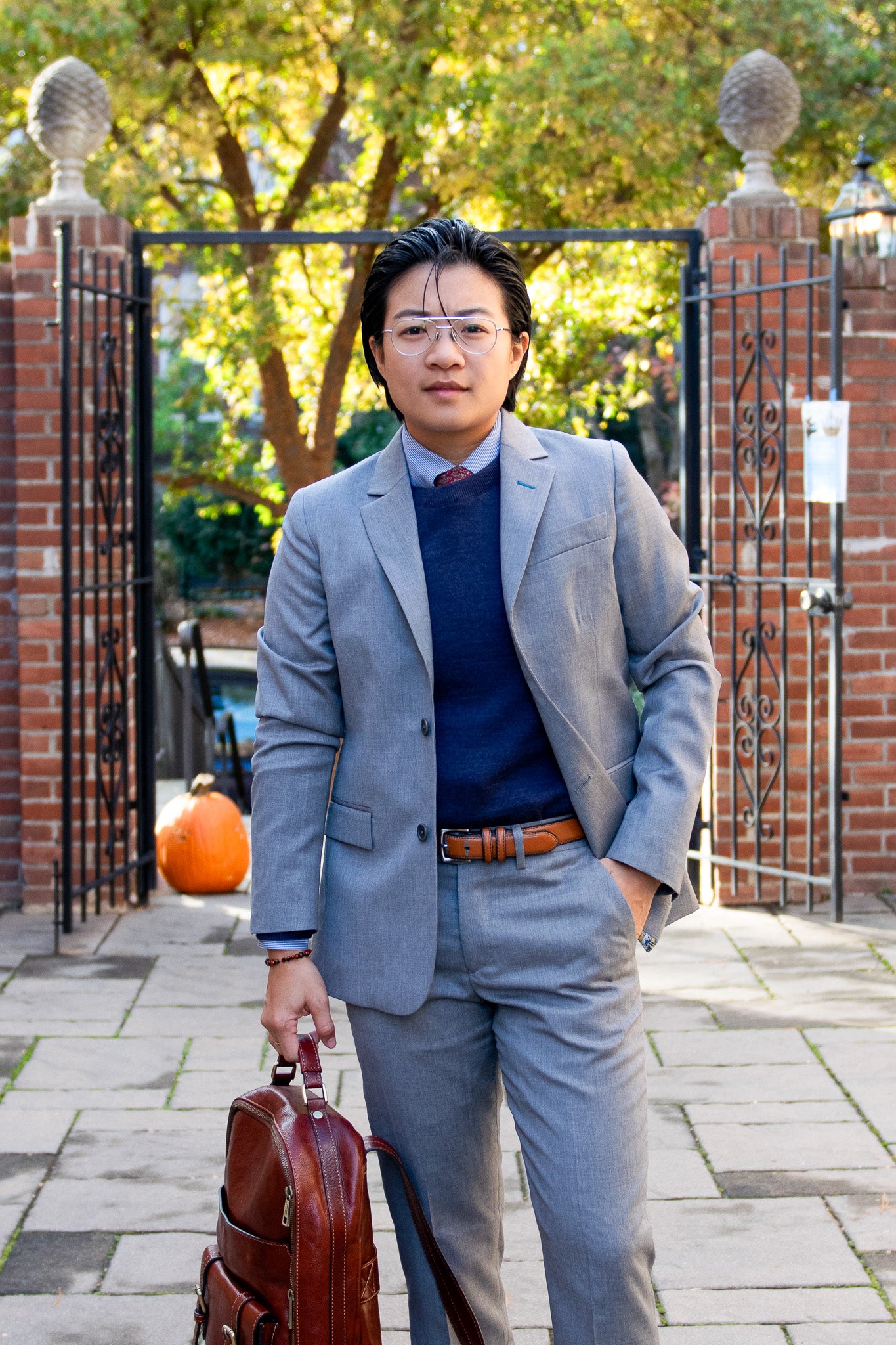 Airin Yung, transmasculine lawyer stands outside in their courtyard in a Kirrin Finch Gray Georgie Suit, Navy Crew Sweater, holding a bag in their left hand. Their hair is slicked back and they dawn a pair of glasses