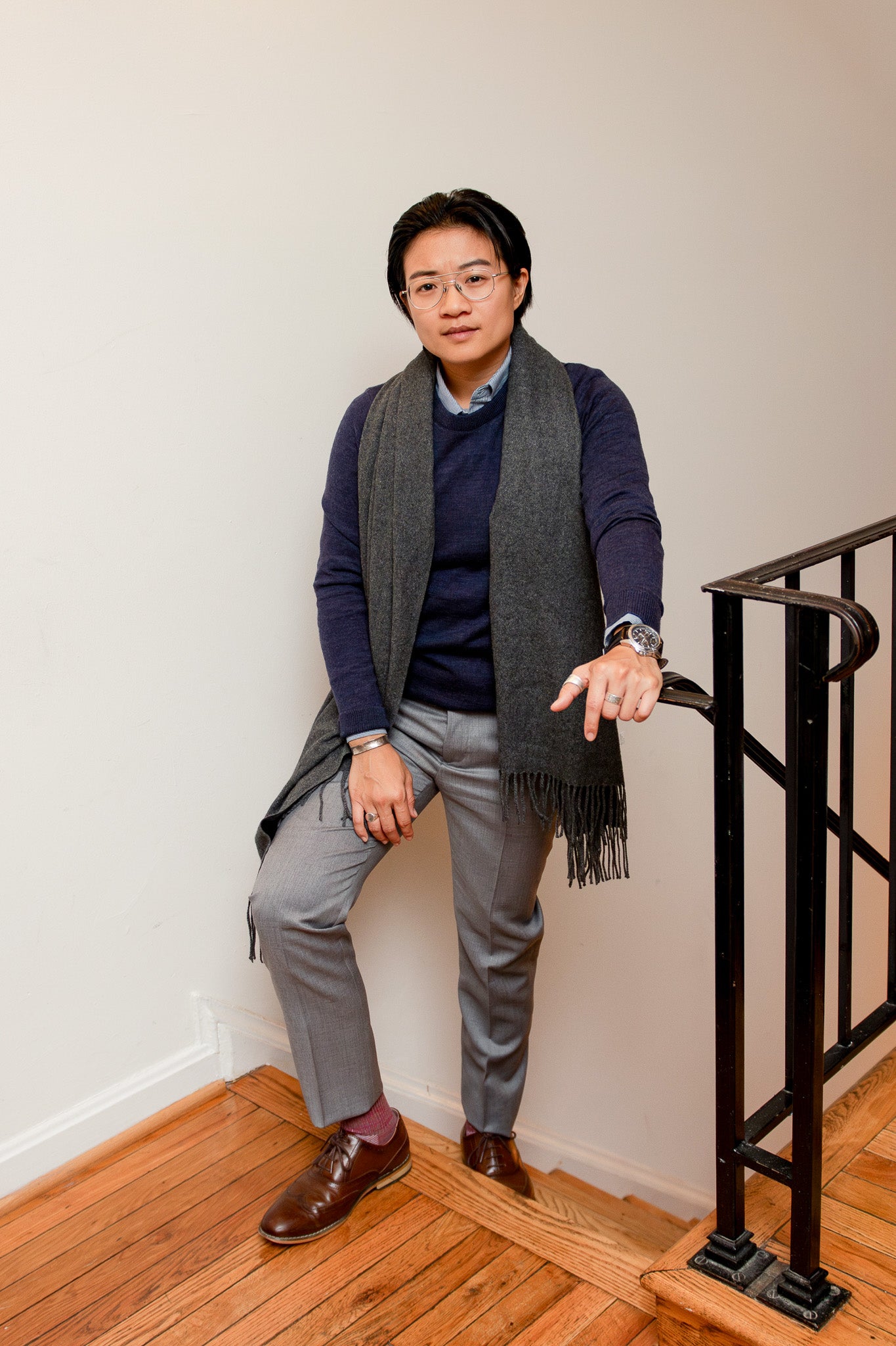 Airin Yung, transmasculine lawyer, stands at the top of the stairs in their home in a Navy Kirrin FInch Merino Wool Sweater, Kirrin Finch Gray Georgie Pants, Kirrin Finch Blue Mini Stripe Dress Shirt, and a gray scarf laid around their neck