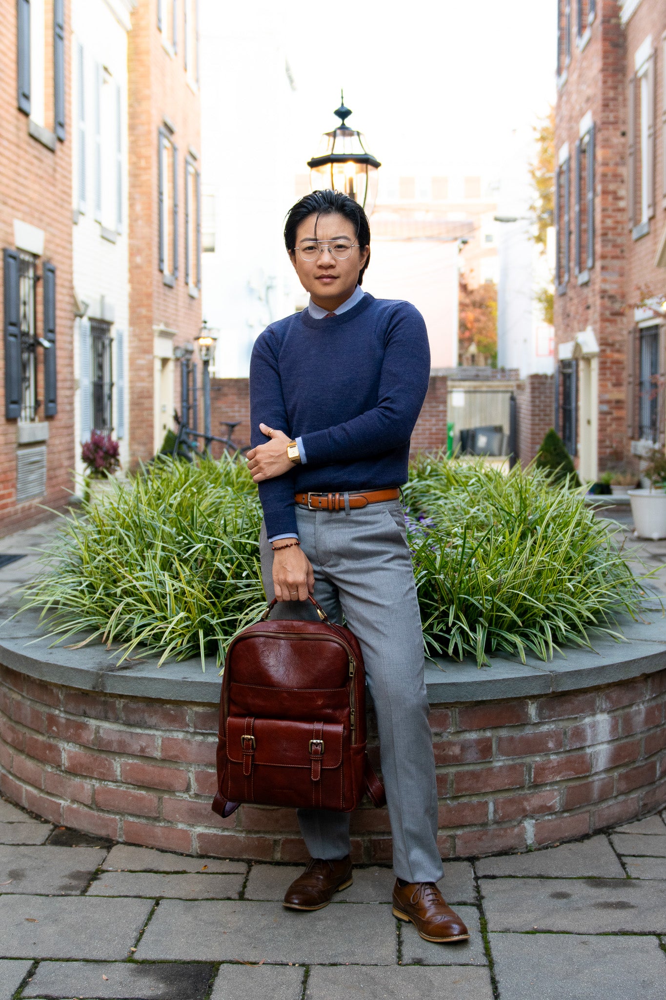 Airin Yung, transmasculine attorney, stands in their outdoor courtyard in a navy blue crew sweater, gray Kirrin Finch Georgie pants, and a blue mini stripe shirt