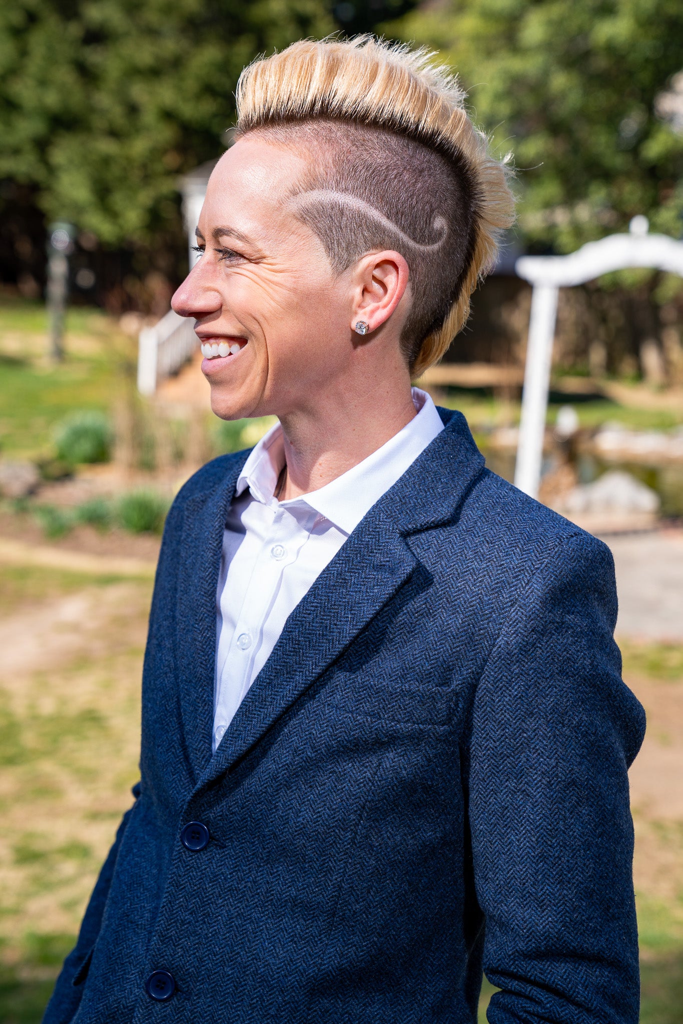 Jo Lohman Standing Sideways in a dark navy Tweed Blazer and white button down. Her Hair is blonde and cut into a mohawk shape with a curvy design line on the side of her head