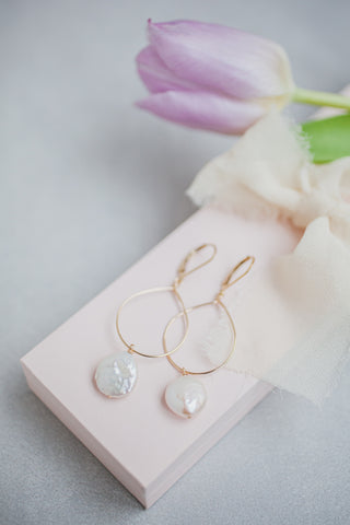 Styled photo of gold hoop earrings with large white freshwater coin pearl drops by J'Adorn Designs jewelry artist Alison Jefferies. Earrings are on a pink jewelry box with a white silk ribbon and purple tulips.