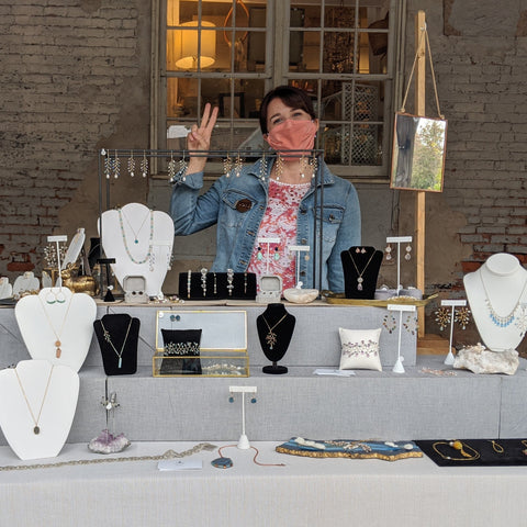 Jewelry artist Alison Jefferies is smiling in a mask and making a peace sign from behind her craft market booth, selling her handcrafted gemstone jewelry and heirloom quality hair accessories.