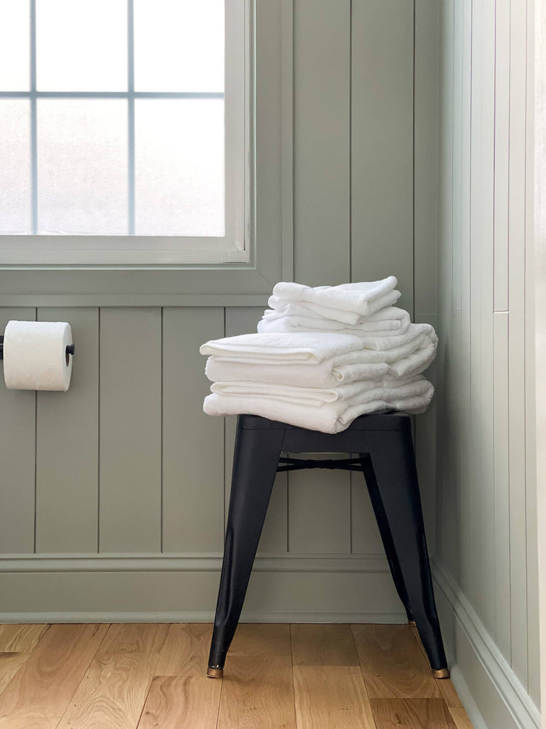 Authenticity50 Essential Cotton Bath Towels in White, stacked on a black stool in the corner of a bathroom with green walls
