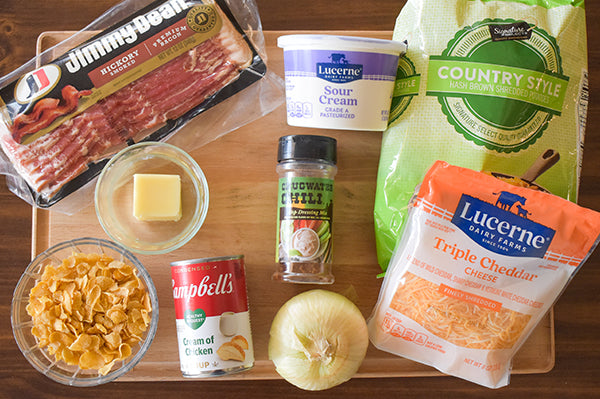 ingredients for loaded cheesy hashbrowns bacon, cream of chicken soup, sour cream, frozen hashbrowns, cheddar cheese, onion, cornflakes, butter, and chugwater chili dip mix