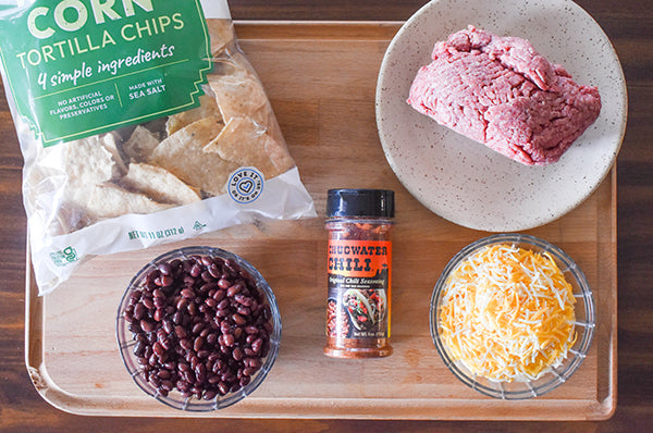 ingredients for sheet pan beefy nachoes ground beef, shredded cheese, black beans, chugwater chili seasoning, and tortilla chips