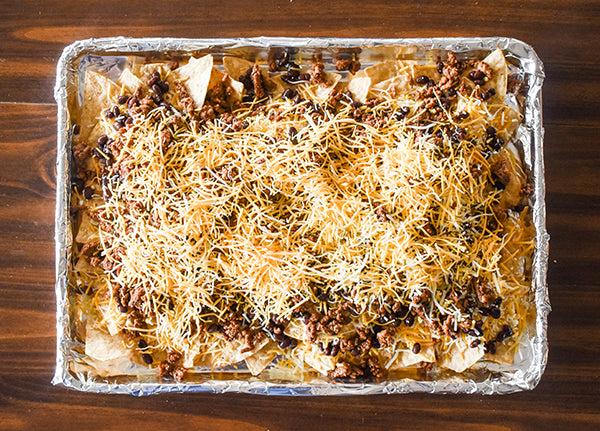 more shredded cheese added on top of tortilla chips covered in cheese, black beans, and ground beef