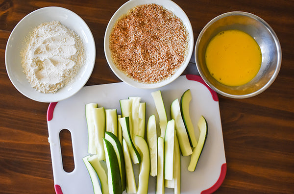 zucchini ready to be breaded, with flour, egg mixture, and seasoned panko crumbs on separate plates 