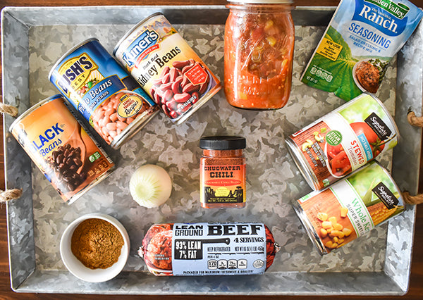 ingredients on tray for taco soup, jar of salsa, can of pinto beans, black beans, kidney beans, ranch dressing mix, ground beef, chugwater chili seasoning, onion, cumin, kernel corn, and stewed tomatoes