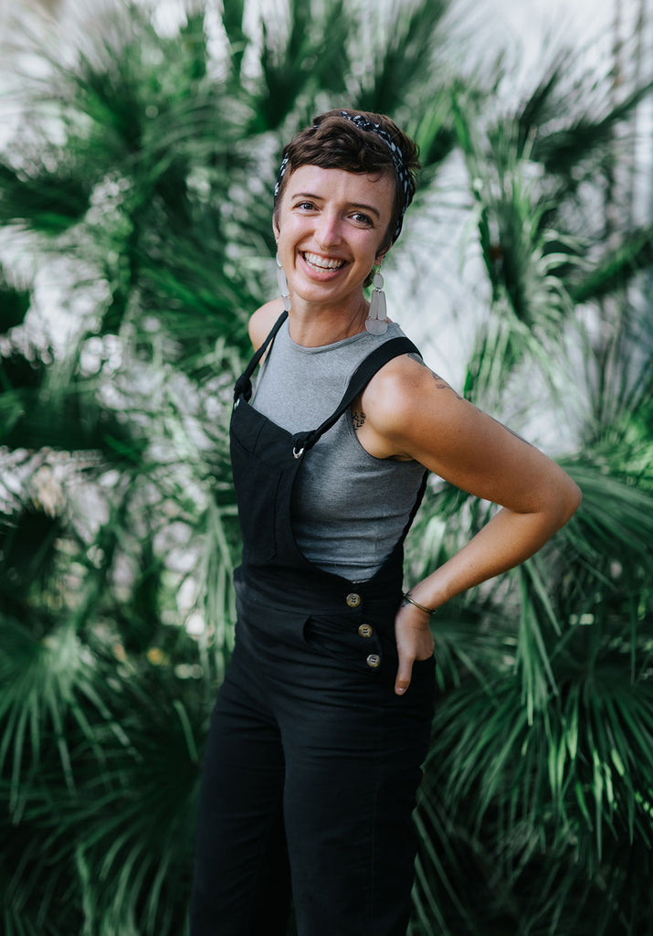 Jess is wearing the RJ Overalls in black over the Ribbed Crop Top in grey in front of a large leafy plant.