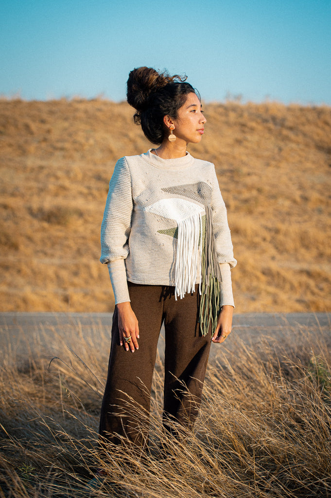 Kristy wearing an olive and green handwoven fringe sweater in front of a blue sky and yellow grasses