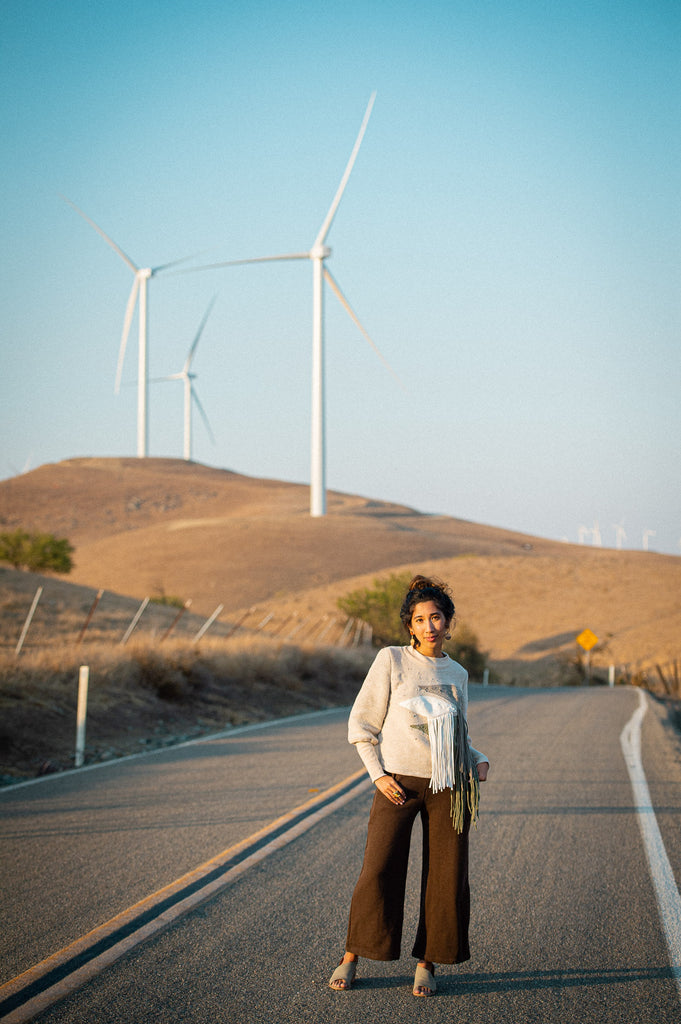 Kristy standing in front of wind turbines wearing a fringe sweater and chocolate brown pants