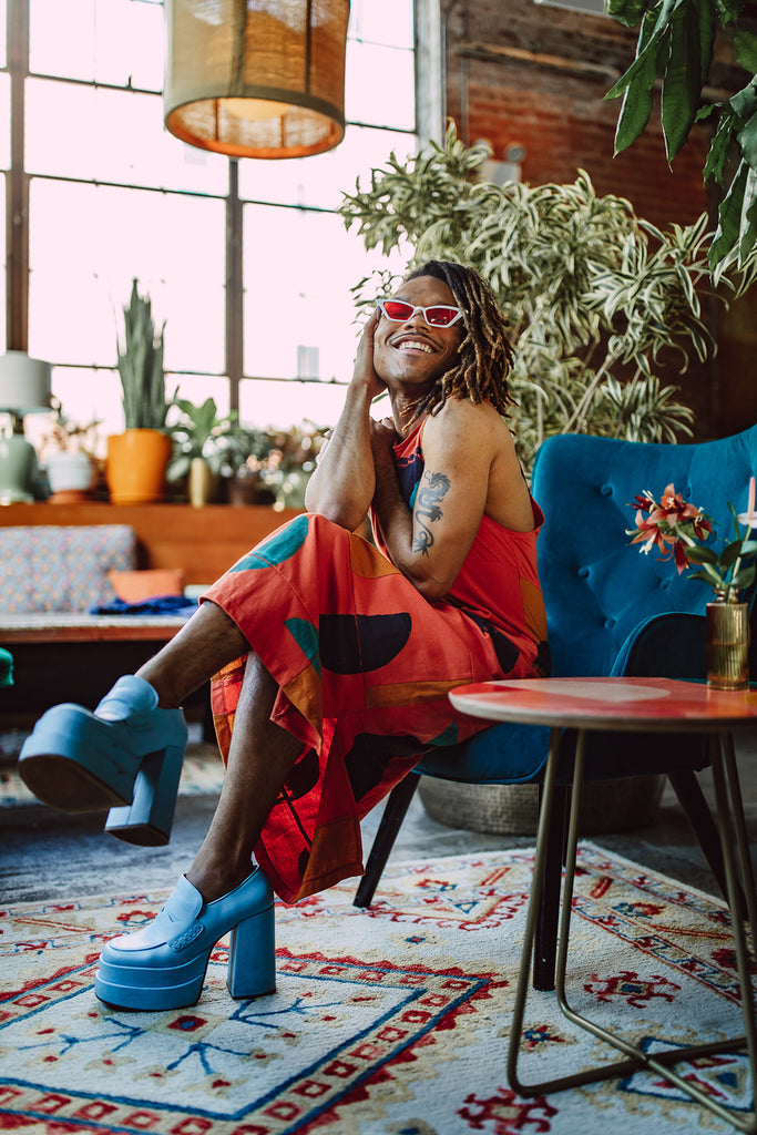 Christopher wears the patchwork anja dress in orange from the plant kween x tonlé collection paired with light blue platform shoes. 