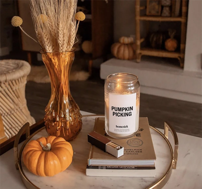 a homesick pumpkin picking candle next to a small pumpkin and a vase of dried wheat