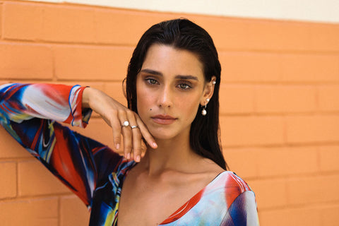 Brunette woman poses against an orange brick wall, wearing pearl earrings, a pearl ring, pearl ear cuff and a colourful dress