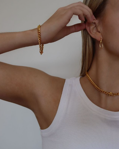 close up of womans arm wearing a gold chain bracelet, with a gold chain necklace around her neck