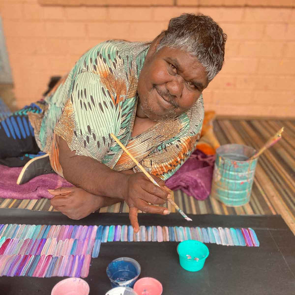 Jeannies Mills Pwerle leaning over a painting in progress, looking at the camera and smiling. She is holding a paintbrush and her blouse and blanket match the colours in the painting.