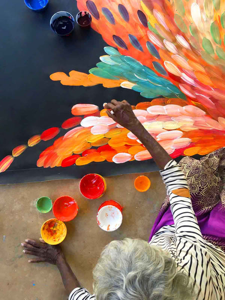 Birds eye view of Gloria Petyarre sitting on porch painting a large orange leaves painting