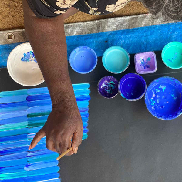 Jeannie Mills' right arm stretches across a black canvas laying on the ground. She is holding a paint brush gracefully. Several paint pots are grouped on the dry canvas and half the painting has disjointed stripes in mostly blue tones painted on it. 
