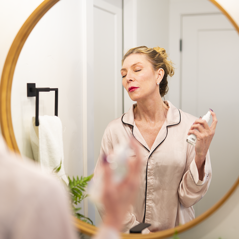 A white woman with blonde hair wearing cream colored pajamas puts on skincare in front of a round mirror