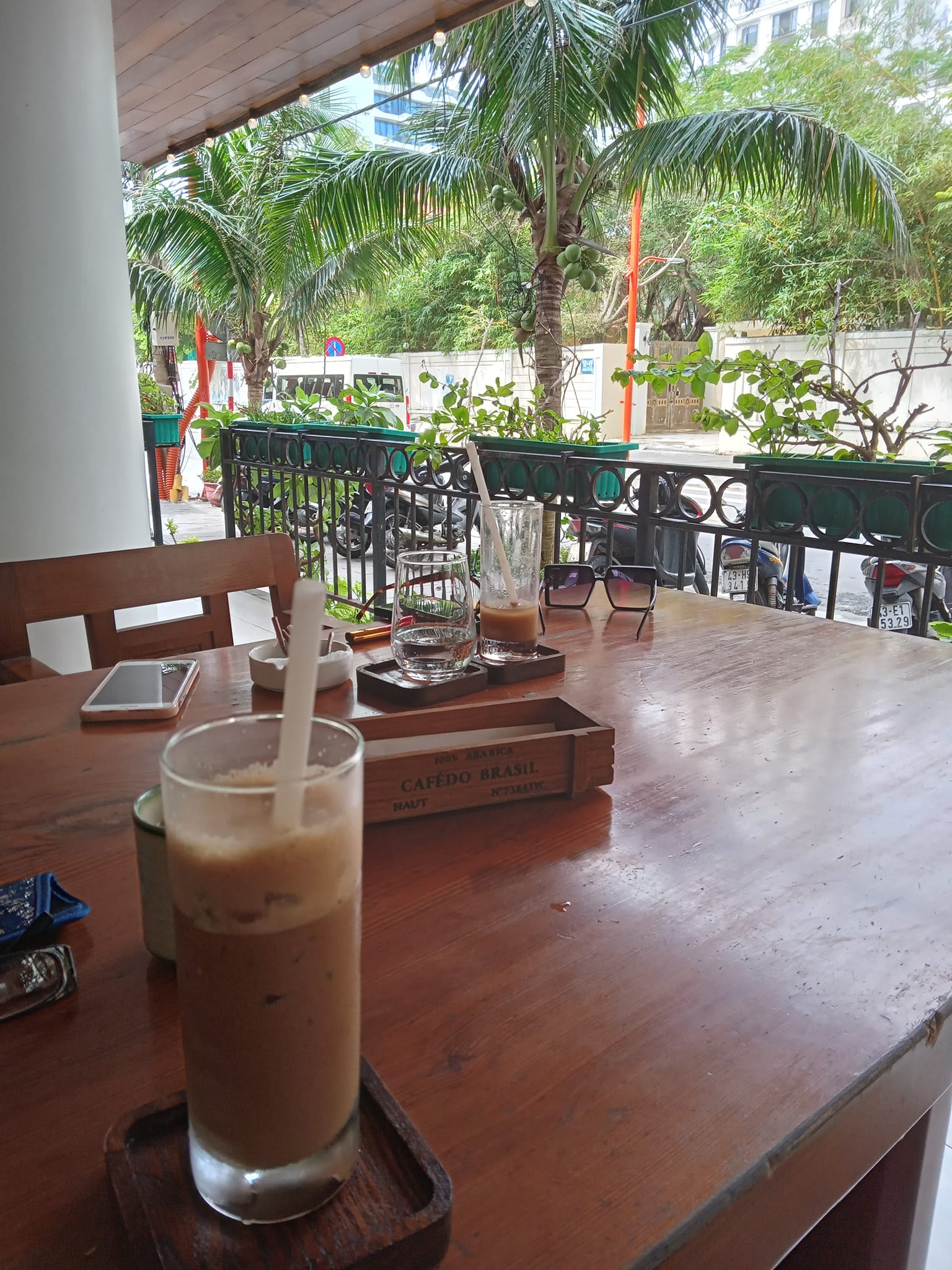 A Vietnamese iced coffee drink in a tall glass with a straw, on the table of an open air cafe in Vietnam, with lush palm fronds lining the open walls