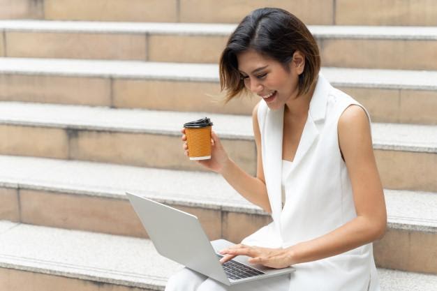 Woman sitting outdoors on steps with laptop on her lap and coffee in hand, smiling