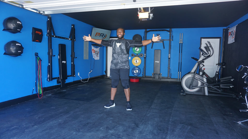 Eric C. standing in the middle of his garage gym that is painted blue, outfitted with PRx Performance racks, weights, benches, wall balls.