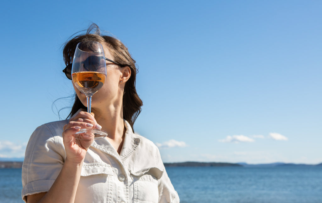 A woman in a white blouse in front of a blue lake holds a glass of pink wine in front of her face.