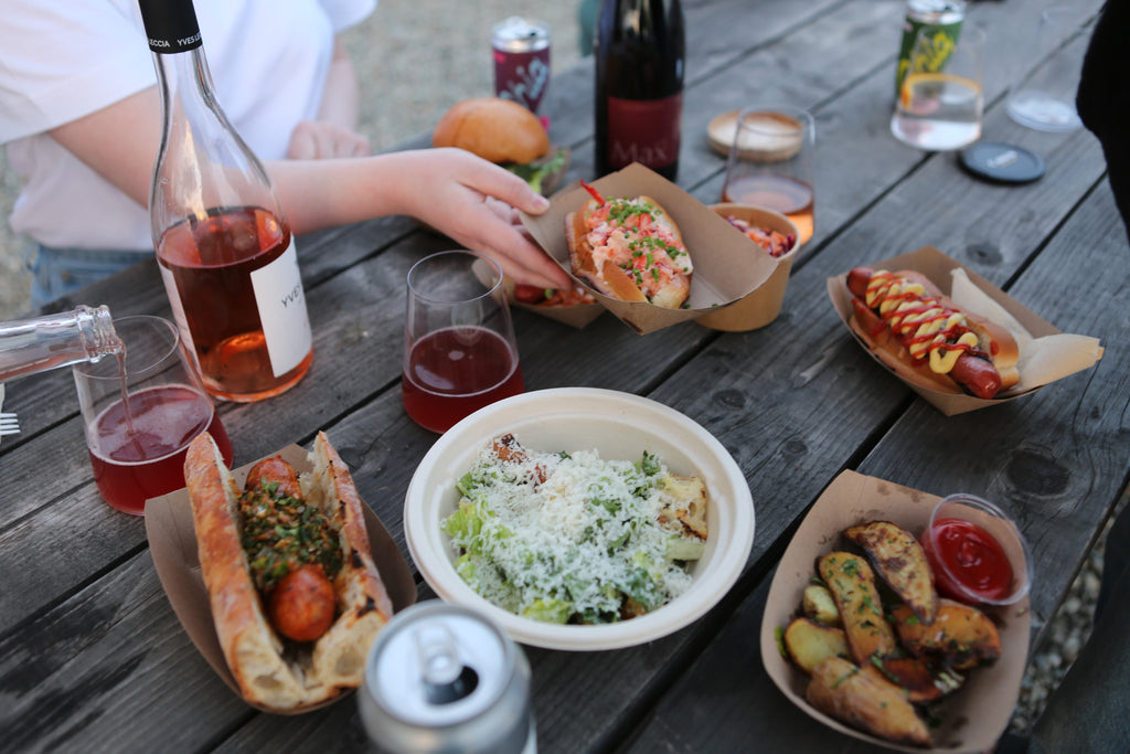 A picnic table loaded with hot dogs, wedge fries, Caesar salad and dark pink wine in stemless glasses.