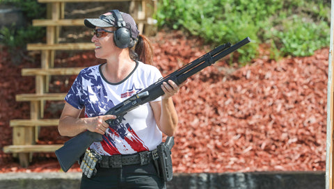 A smiling woman in a cap and earmuffs aims a handgun, wearing slim, protective sunglasses.