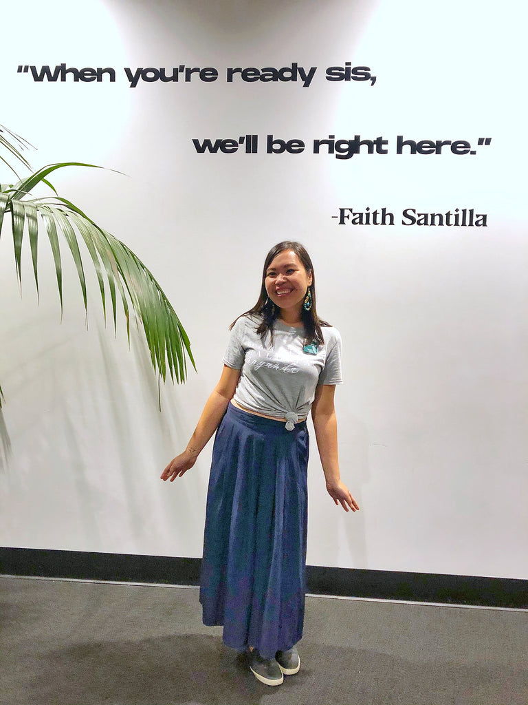 Gelaine Santiago, Social Entrepreneur and Co-Founder of Cambio & Co., standing beneath a sign that says 'when you're ready sis, we'll be right here' by Faith Santilla, at the Entrepinays Summit 2019 in San Francisco