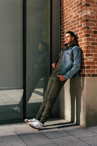 Tall man leaning against brick wall wearing jean jacket over camo green sweatsuit