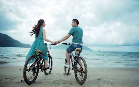 a couple wearing calming green colors bicycling on the beach as they hold hands