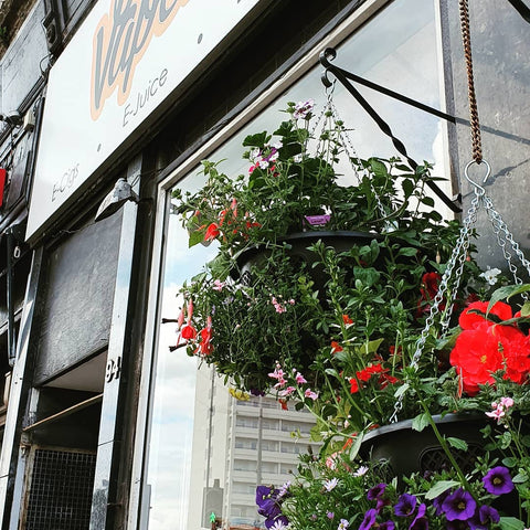 Shows the outside of the Vapetek shop in spring. Also in view is a hanging basket filled with flowers.